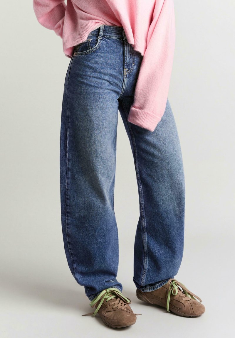 Personne portant un jean large bleu, un pull oversize rose et des baskets marron avec des lacets verts, debout devant un fond uni.