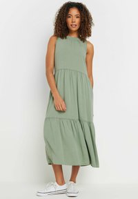 Ljusgrön ärmlös maxiklänning med lager av volanger, en samlad halsringning och mjukt bomullstyg, i kombination med vita sneakers.
