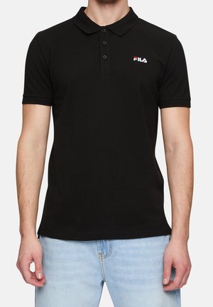 Mężczyzna w czarnej, krótkorękawowej koszulce polo z logo FILA na piersi i jasnoniebieskich dżinsach, stoi na jednolitym tle.