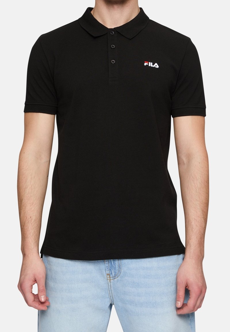 Homme portant un polo noir à manches courtes avec le logo FILA sur la poitrine et un jean bleu clair, debout devant un fond uni.