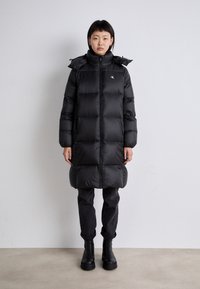 Črn puffer plašč z visokim ovratnikom, sprednjim zadrgo in kapuco. Ima prešito vzorčenje, dolgo dolžino in stranske žepke. Nosite ga preko temnih hlač.