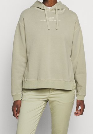 Osoba oblečená v světle béžové mikině s kapucí a šňůrkami a bílým nápisem na hrudi, doplněná o odpovídající světle béžové kalhoty.