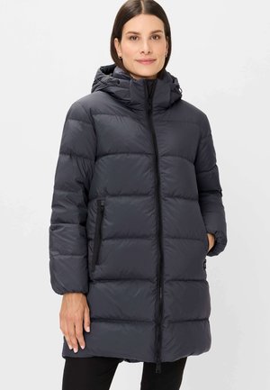 Frau trägt eine lange schwarze Daunenjacke mit Kapuze, Reißverschluss vorne und Seitentaschen, steht vor einem einfarbigen hellen Hintergrund.