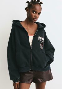 Čierna mikina na zips so vyšitým štítkom s nápisom "FREEDOM DON'T" a dizajnom shield. Nosené cez svetlý tielko, spárované s hnedými šortkami.