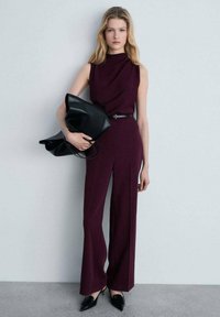 Combinaison sans manches bordeaux avec un col haut, une taille cintrée et une coupe à jambes larges. Assortie à un sac fourre-tout noir et à des talons noirs élégants.