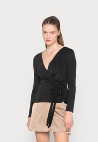 Top preto envolto com mangas longas, decote em V e um laço na cintura. Tecido liso com um brilho sutil. Combinado com uma saia bege.