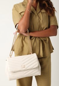 Borsa a tracolla bianca trapuntata con catena, caratterizzata da un accento dorato a forma di M. Design strutturato con chiusura a patta, posata su un completo color khaki.