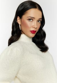 Witte, fluffy trui met hoge hals en textuurstof. Model draagt hangende pareloorbellen en gedurfde rode lippenstift. Neutrale achtergrond.