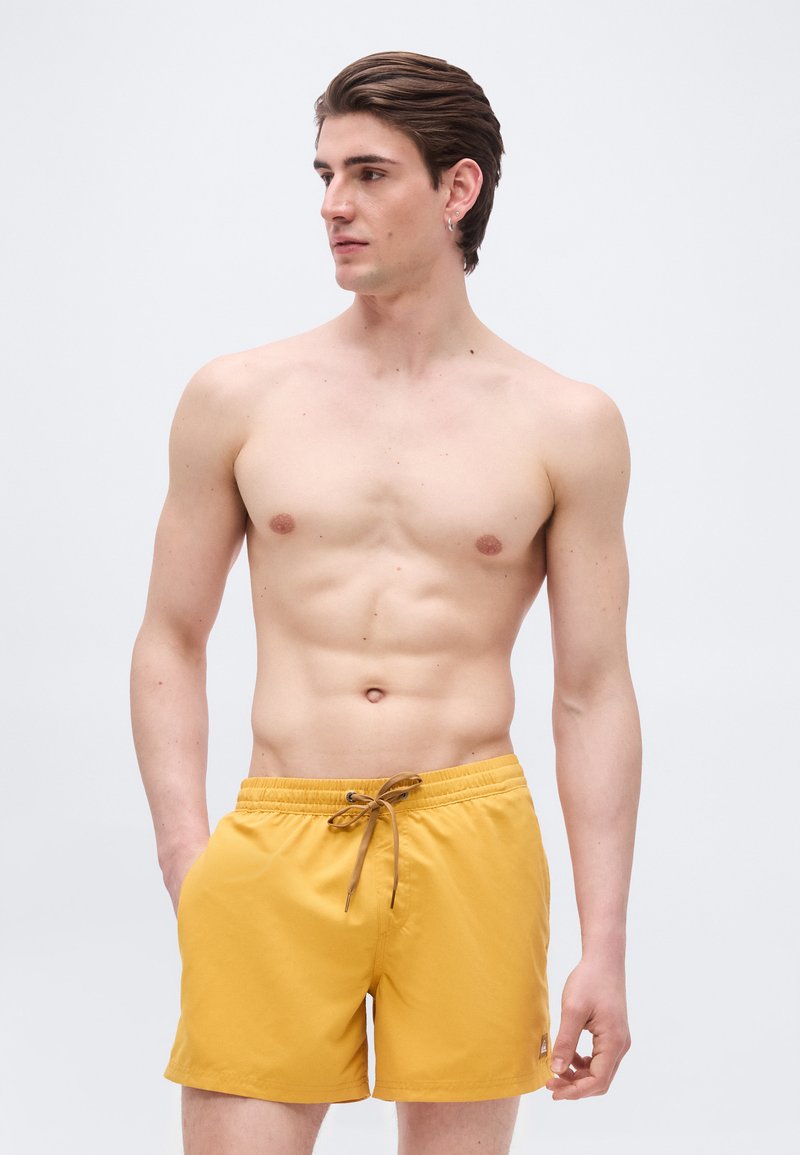 Νέος άνδρας με κοντά καστανά μαλλιά, που φοράει κίτρινα μαγιό, στέκεται με το ένα χέρι στην τσέπη, κοιτάζοντας προς το πλάι σε απλό φόντο.