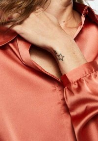 Satin orange bluse med krave, knaplukning og elastiske manchetter. En håndledstatovering af en stjerne er synlig på venstre håndled.