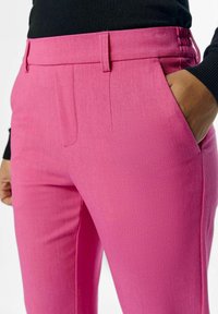 Pantalons ajustés roses en tissu texturé, présentant un devant plat avec des poches latérales et des passants de ceinture, conçus pour une silhouette cintrée.