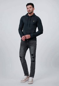 Svart textured hettegenser med vertikal ribbestruktur, gull logo på brystet, kombinert med slitte grå jeans og hvite sneakers.
