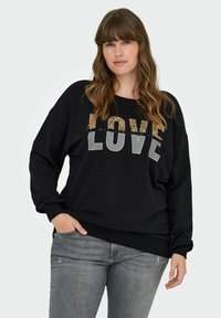 Sieviete ar gariem viļņainiem matiem, tērpusies melnā džemperī ar uzrakstu "LOVE" leoparda izdrukā un pelēkos burtos, un pelēkās biksēs, pozējot ar roku kabatā.