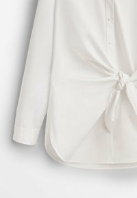 Chemise blanche à boutons avec manches longues, présentant un détail noué à la taille et un ourlet courbé. Tissu lisse avec une légère brillance.