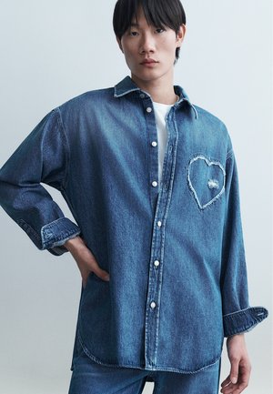 Noor inimene, kes kannab sinist denimist nööbitavat särki, millel on rindkere piirkonnas õmblusteta südamekujuline muster, sobitatud samast denimmaterjalist pükstega.