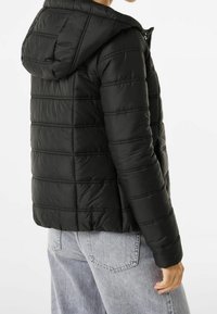 Černá péřová bunda s kapucí, se švy ve tvaru kosočtverce, hladkým povrchem a zipovým uzávěrem. Nositelná přes světle modré džíny.