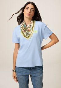 Camiseta de algodón azul claro con cuello redondo, que presenta un gráfico en amarillo neón. Combinada con una bufanda de seda estampada alrededor del cuello.