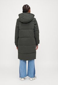 Lang, mørkegrønn pufferjakke med hette, med horisontale quiltede seksjoner, kombinert med vide blå jeans og hvite sko.