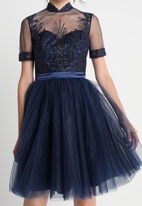 Abito blu navy con corpino aderente caratterizzato da dettagli intricati di perline, maniche corte in chiffon e gonna flottante in tulle. Dettaglio in satin in vita.