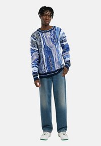 Gestrickter Pullover in Blautönen mit gemustertem Design, kombiniert mit locker sitzenden hellblauen Jeans und weißen Sneakers mit grünen Akzenten.