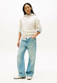 Beige Sweatshirt mit Kapuze, blaue karierte Hemd darunter, hellblaue weite Jeans und weiße Loafers mit Stickdetails.