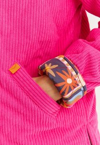 Polarowy pulower w jasnoróżowym kolorze o prążkowanej teksturze, z kolorowym kwiatowym wzorem na mankietach i małą pomarańczową metką z boku.