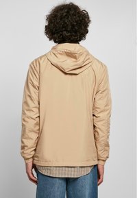 Beige hættetrøje med en glat, let skinnende tekstur, elastiske manchetter og en afslappet pasform, lagdelt over en ternet skjorte og jeans.