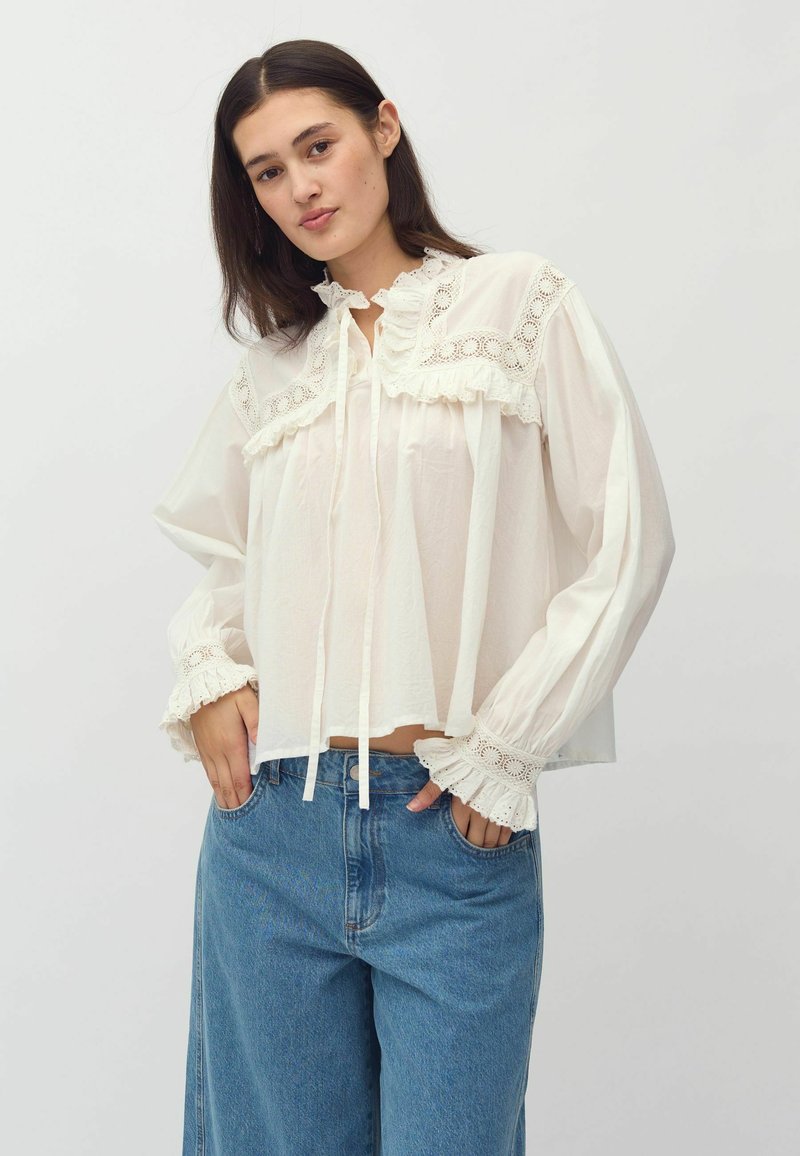 White blouse with lace trim and ruffled collar; long sleeves with lace cuffs. Paired with casual blue jeans. Soft, lightweight fabric.