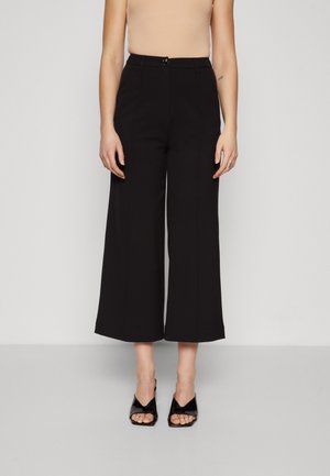 Woman wearing high-waisted black wide-leg cropped pants and black open-toe heeled sandals, standing against a plain light background.