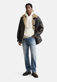 Black shearling-lined coat over a white hoodie, paired with light blue denim jeans and dark shoes. The outfit features contrasting textures.