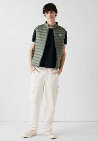 Khaki pufferbodywarmer over een donkergroen T-shirt, gecombineerd met witte cargobroek en crèmekleurige sneakers; heeft een hoge kraag en een logopatch.