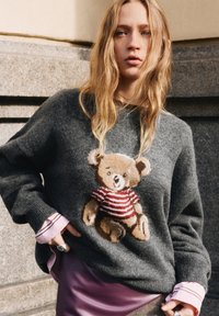 Szary dzianinowy sweter z haftowanym misiem w czerwono-białą paskowaną koszulką. Długie rękawy z prążkowanymi mankietami, noszony na różowej koszuli.