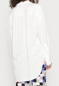 Vit långärmad skjorta med en veck bak, rundad nederkant och knappmanschetter, gjord av ett slätt tyg. Kombinerad med flerfärgade rutiga byxor.