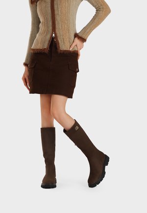 Donna con gonna di velluto a coste marrone, cardigan beige lavorato a maglia con bordatura in pelliccia e stivali al ginocchio marrone scuro su sfondo uniforme.