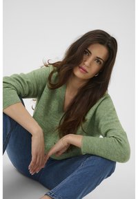 Pull vert à col en V en maille douce et texturée. Porté avec un jean bleu, le modèle est assis confortablement, mettant en valeur la coupe décontractée et les poignets côtelés.