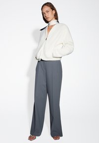 Giacca bomber bianca e fluffy con zip e tasche laterali, abbinata a pantaloni grigi a vita alta. L'outfit è completato da una sciarpa bianca e nera.