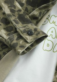 Chemise à motif camouflage dans des tons olive et noir, avec une manchette boutonnée et une chemise blanche en dessous avec du texte vert. Tissu texturé visible.