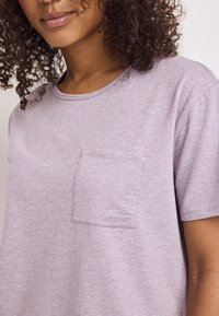 Camiseta de color púrpura claro con cuello redondo, mangas cortas y un bolsillo en el pecho con un discreto bordado de la marca. Tejido suave y texturizado.