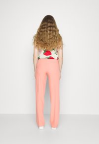 Persoon met lang golvend haar, gekleed in een witte mouwloze top met aardbeienprint en een hoge taille roze broek, kijkt van zich af op een effen achtergrond.