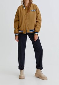 Osoba nosząca brązową kurtkę typu varsity z napisem "1991" na piersi, czarne spodnie z podwiniętymi nogawkami oraz beżowe masywne buty, stojąca na jednolitym tle.