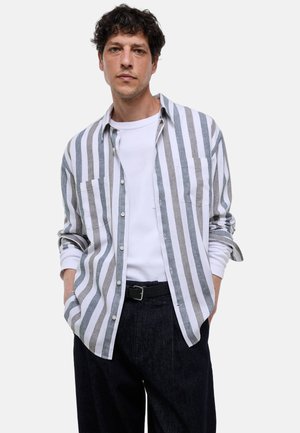 Homme portant une chemise à boutons rayée verticalement gris et blanc sur un t-shirt blanc à manches longues, ceinture noire et pantalon foncé, mains dans les poches.