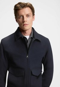 Dark navy fleece jacket with a zippered front, two front pockets, and a pointed collar, worn over a gray shirt. Smooth texture.