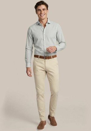 Lachende man die een groen-wit gestreept hemd met knopen, beige broek, bruine riem en bruine instapschoenen draagt, staand voor een effen achtergrond.