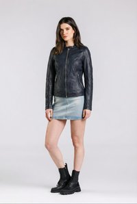 Nainen seisoo musta nahkatakki päällään, vaaleansininen farkkuminihame ja mustat nyörättävät maiharit jalassa yksiväristä taustaa vasten.