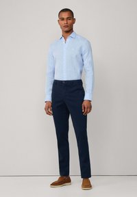 Camicia azzurra chiara a maniche lunghe con bottoni, realizzata in tessuto testurizzato, abbinata a chinos blu navy e mocassini marroni, in piedi su uno sfondo neutro.