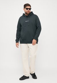 Felpa verde scuro con tasca frontale, cappuccio con cordino e dettaglio del logo. Abbinata a pantaloni beige chiaro e sneakers nere.