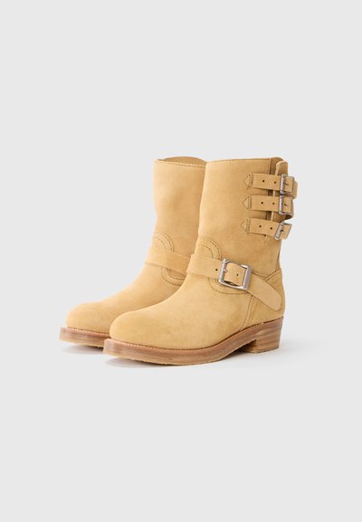 Botas de ante amarillas hasta el tobillo con punta redonda, tres correas laterales y hebillas plateadas. Tacón de aspecto de madera con un acabado suave.