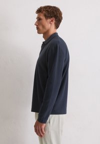 Młody mężczyzna stojący bokiem, ubrany w granatową koszulę z długim rękawem i beżowe spodnie, na tle gładkiej białej ściany.