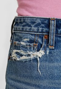 Blaue Jeans aus Denim mit einem abgenutzten Design, das ausgefranste Kanten und sichtbare Nähte aufweist. Kupferfarbene Nietenakzente an der Taille.