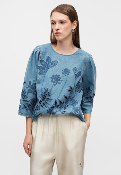 Blouse en jean à manches trois-quarts, ornée d'une broderie florale détaillée et de découpes dans différentes nuances de bleu, avec un col rond.